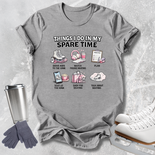 A gray graphic t-shirt laid flat on a light gray textured background, surrounded by ice skating accessories including white ice skates, a silver travel mug, and gray gloves. The t-shirt features a white and pink graphic with the text 'THINGS I DO IN MY SPARE TIME' and six small illustrations with labels.