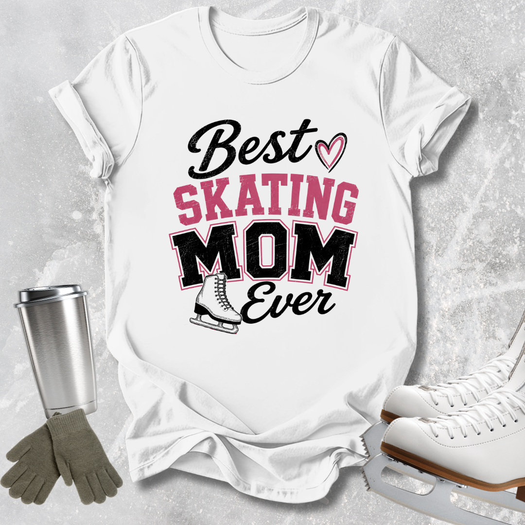 A white graphic T-shirt laid flat on a light gray textured surface, with a silver travel mug, gray gloves, and white ice skates around it. The shirt reads 'Best SKATING MOM Ever' in black and pink lettering with a small heart and skater boot graphic.