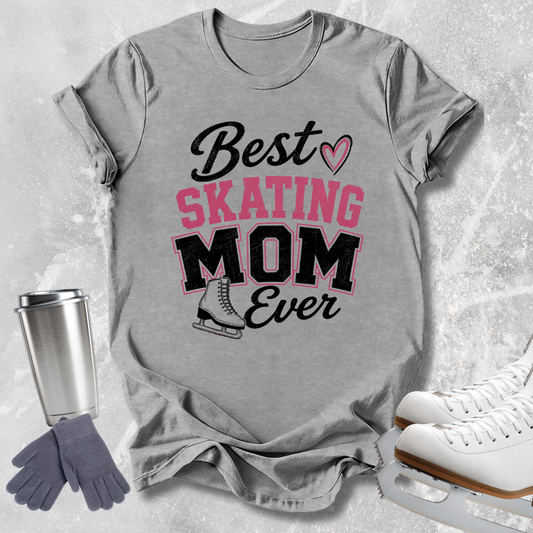 A heather gray graphic t-shirt laid flat on a light gray textured background, featuring the text 'Best SKATING MOM Ever' in black and pink with a small ice skate illustration. Surrounding props include a stainless steel travel mug, a pair of gray gloves, and white ice skates.