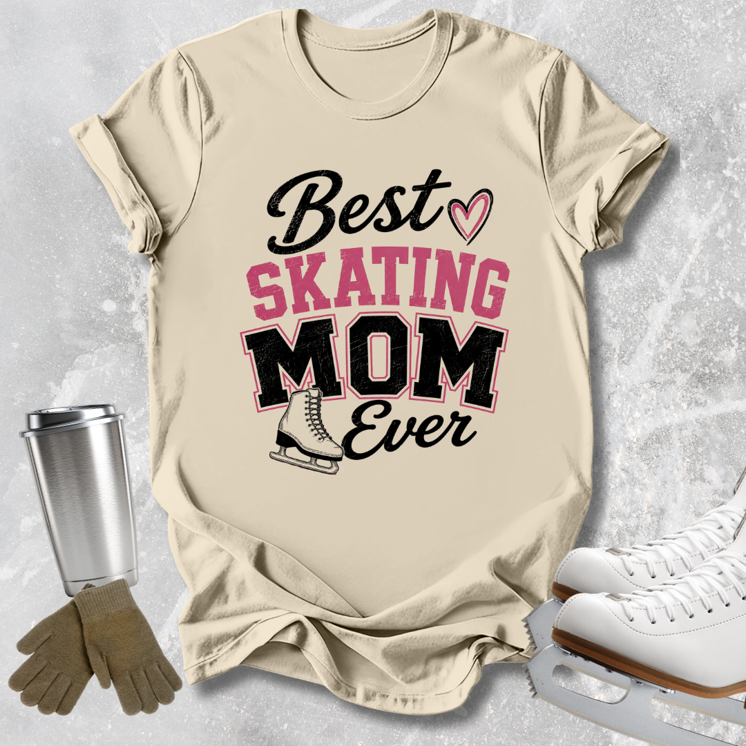 A beige graphic T-shirt laid flat on a light gray textured surface, with skating-themed props around it: a stainless steel cup, gloves, and ice skates. The shirt reads “Best SKATING MOM Ever” in black and pink lettering with a small heart and ice skate illustration.