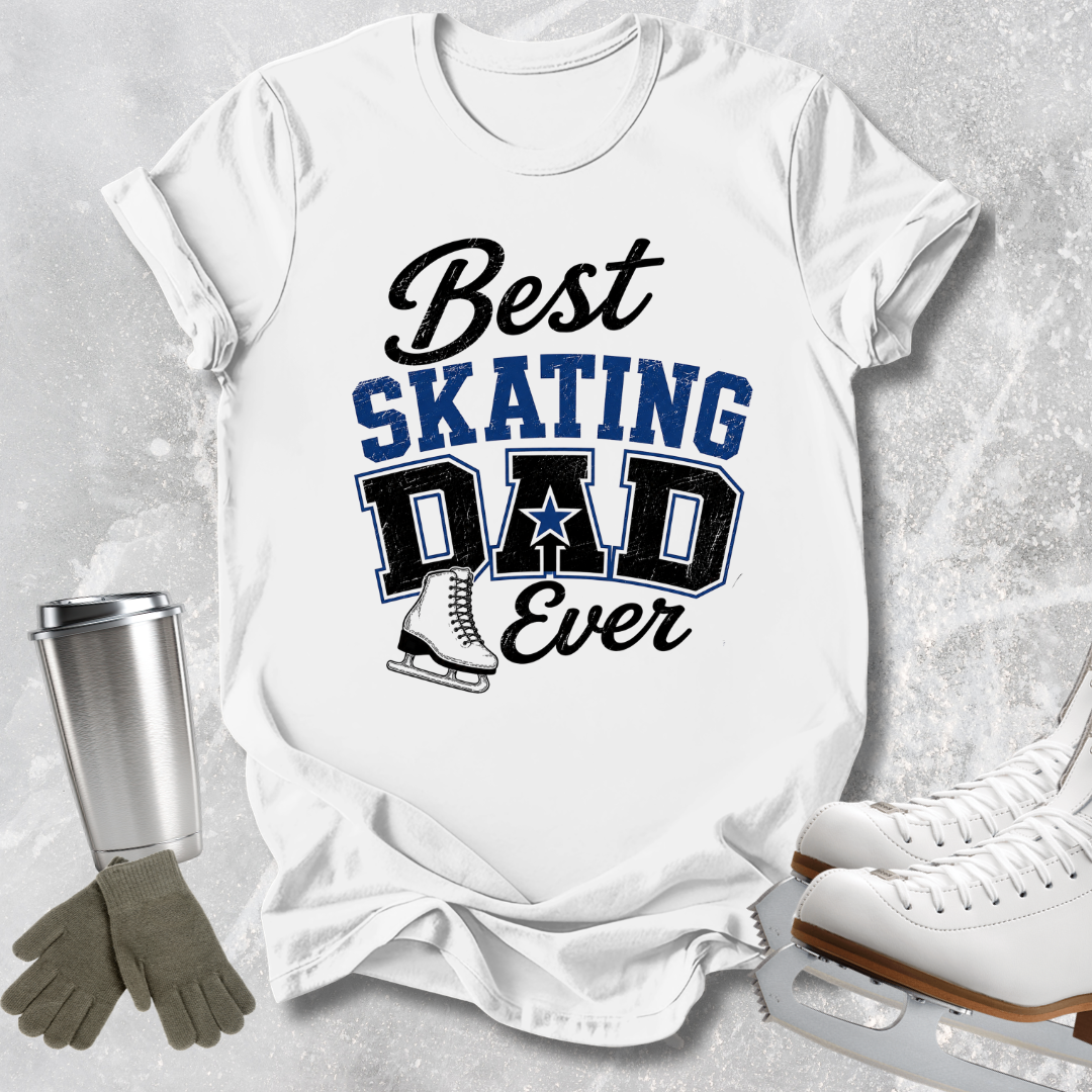 A white graphic t-shirt laid flat on a light gray textured surface, featuring the text 'Best SKATING DAD Ever' in black and blue lettering with a skater boot graphic. Ice skates, gloves, and a metal tumbler are arranged around the shirt as styling props.