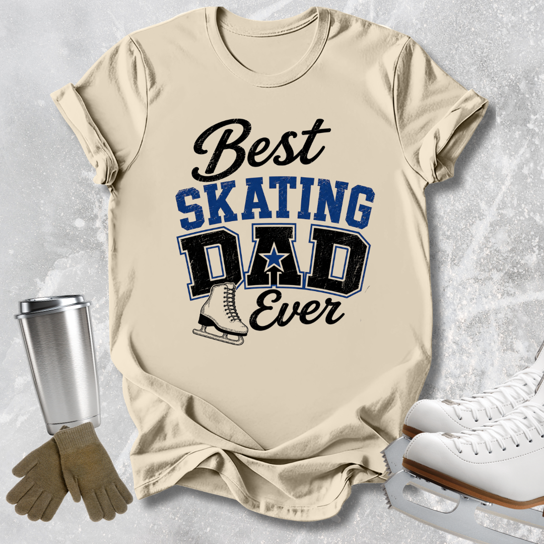 A beige graphic T-shirt laid flat on a light gray textured background, with the text 'Best SKATING DAD Ever' in black and blue lettering and a small ice skate illustration. Ice skates, gloves, and a metal cup are placed around the shirt as styling props.