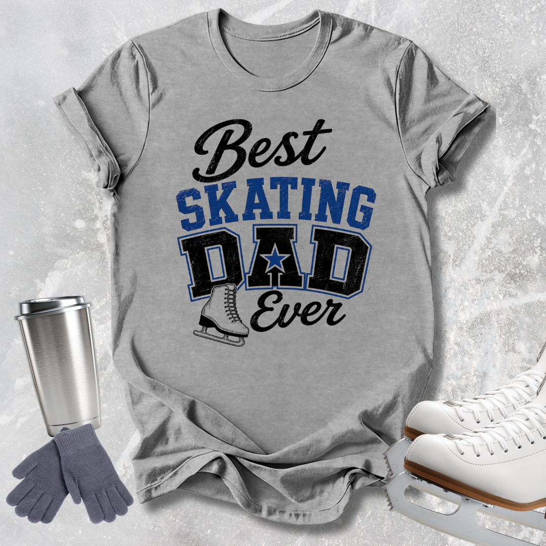 A heather gray graphic t-shirt laid flat on a light gray textured background, featuring the text 'Best SKATING DAD Ever' in black and blue with a skater boot graphic. Surrounding the shirt are ice skates, a silver tumbler, and a pair of gray gloves.