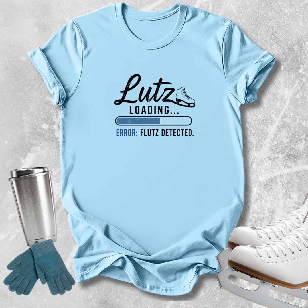 A light blue graphic T-shirt laid flat on a light gray textured surface, with a stainless steel tumbler, teal gloves, and white ice skates around it. The shirt has a black printed design reading 'Lutz Loading.