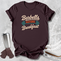 A dark brown graphic t-shirt laid flat on a light gray textured surface, styled with a stainless steel travel mug, brown gloves, and white ice skates. The t-shirt features the text 'Barbells BEFORE Breakfast' in beige and orange with a teal barbell graphic.