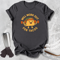 A dark charcoal gray graphic t-shirt laid flat on a light gray textured surface, surrounded by lifestyle props including a stainless steel travel mug, black gloves, and white ice skates. The t-shirt features a cartoon taco character lifting a barbell with the text 'WILL WORK OUT FOR TACOS' in yellow.