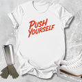 A white short-sleeve T-shirt laid flat on a light gray textured surface, featuring the phrase 'PUSH YOURSELF' in red script across the chest. A stainless steel tumbler, gray gloves, and white ice skates are arranged around it as styling props.
