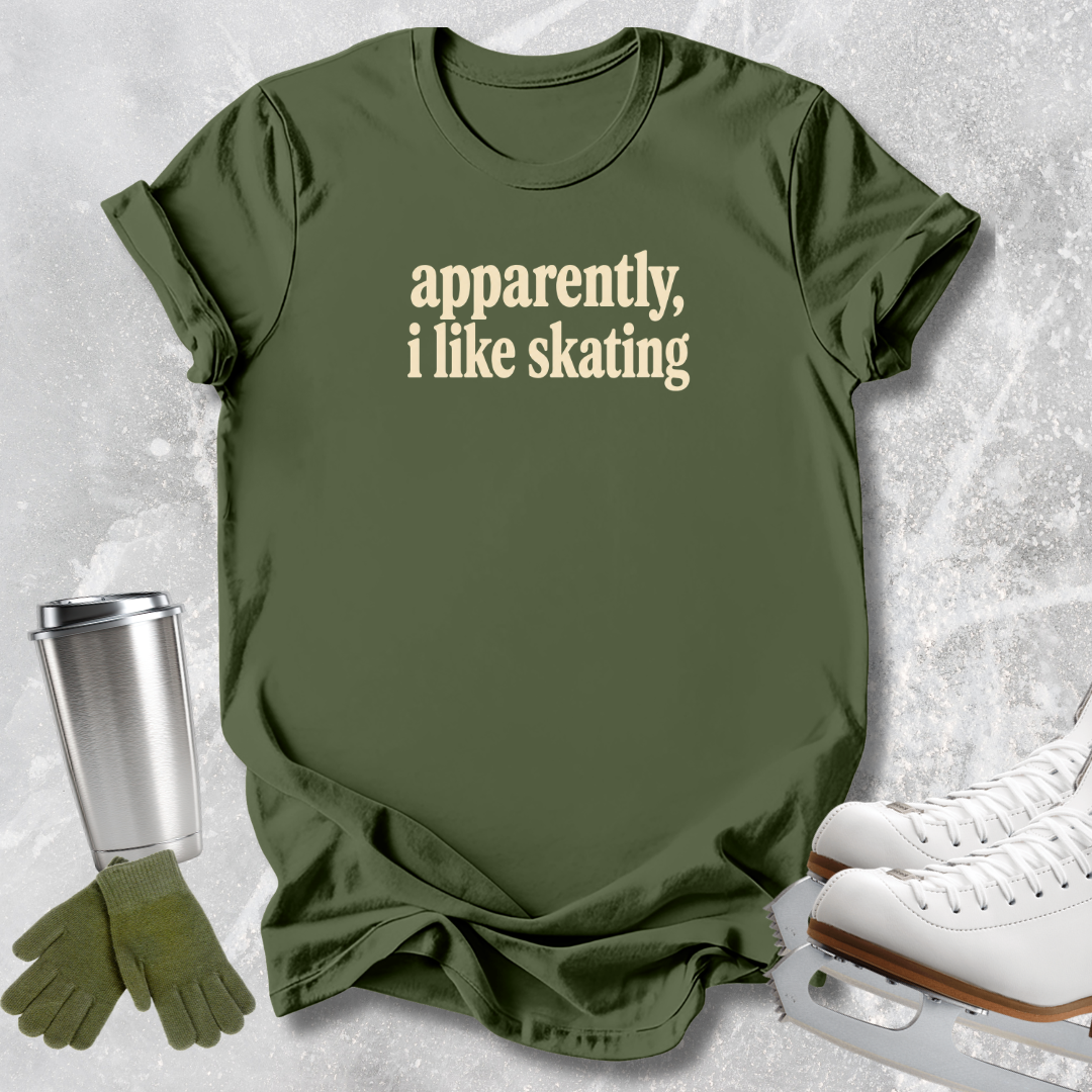 An olive green graphic t-shirt with the text 'apparently, i like skating' in a light beige serif font. The t-shirt is styled in a flat-lay arrangement on a textured gray background, accompanied by white ice skates, a silver travel mug, and green gloves.