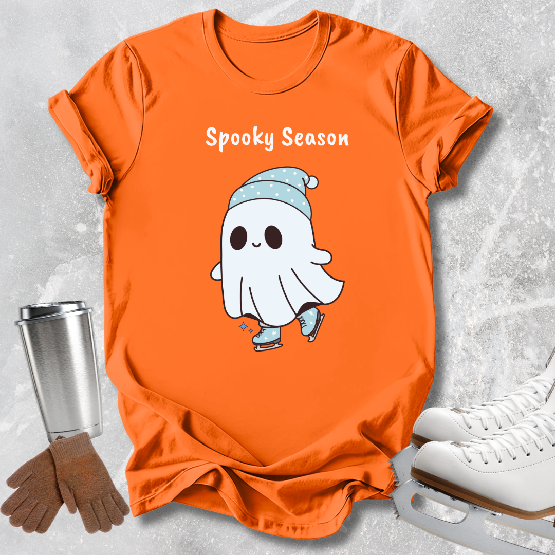 An orange short-sleeve T-shirt laid flat on a light gray textured surface, featuring a white cartoon ghost wearing a light blue hat and ice skates, with the words 'Spooky Season' above it. A stainless steel cup, brown gloves, and white ice skates are placed around the shirt as styling props.