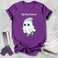 A purple short-sleeve T-shirt laid flat on a light gray textured surface, featuring a white cartoon ghost wearing a light blue hat and skates, with the words 'Spooky Season' above it. A stainless steel travel mug, purple gloves, and white ice skates are arranged around the shirt as props.