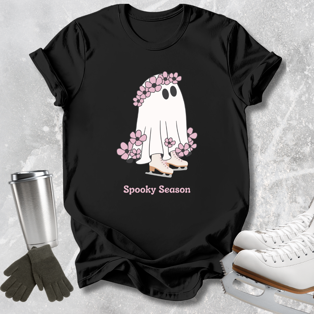 A black short-sleeve graphic T-shirt laid flat on a light gray textured surface, styled with ice skates, gloves, and a metal cup. The shirt features a ghost graphic with pink flowers and the text 'Spooky Season'.