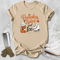 A beige graphic t-shirt laid flat on a light gray textured background, featuring the text 'Pumpkin Spice and Ice' with illustrations of a hot chocolate drink, a pumpkin, autumn leaves, and ice skates. A stainless steel tumbler, gloves, and white ice skates are styled around the shirt.