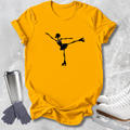 A bright yellow short-sleeve T-shirt laid flat on a light gray textured surface, featuring a black silhouette graphic of a figure skater in a single legged pose. White ice skates, gray gloves, and a silver tumbler are arranged around the shirt as styling props.