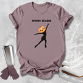 A mauve short-sleeve graphic T-shirt laid flat on a light gray textured surface, with a black silhouette of a figure wearing a jack-o'-lantern head and the words 'SPOOKY SEASON' printed above. A stainless steel tumbler, purple gloves, and white ice skates are arranged around the shirt as styling props.