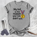 A heather gray graphic t-shirt laid flat on a light gray textured background, surrounded by ice skating props including white ice skates, a silver travel tumbler, and a pair of gray gloves. The t-shirt features the text 'My kid is the reason your kid didn't win.