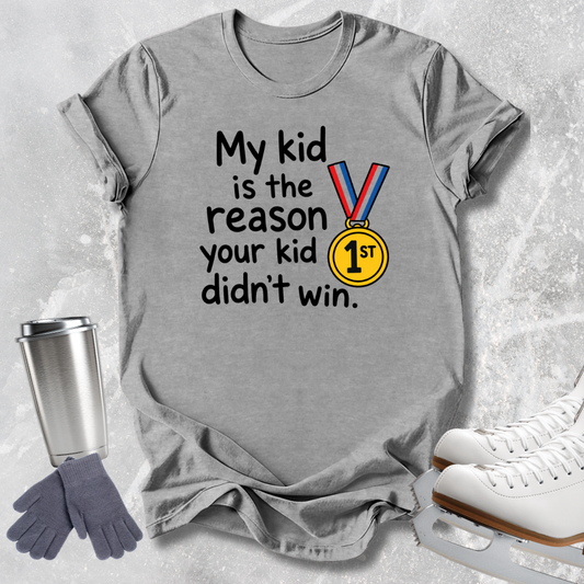 A heather gray graphic t-shirt laid flat on a light gray textured background, surrounded by ice skating props including white ice skates, a silver travel tumbler, and a pair of gray gloves. The t-shirt features the text 'My kid is the reason your kid didn't win.