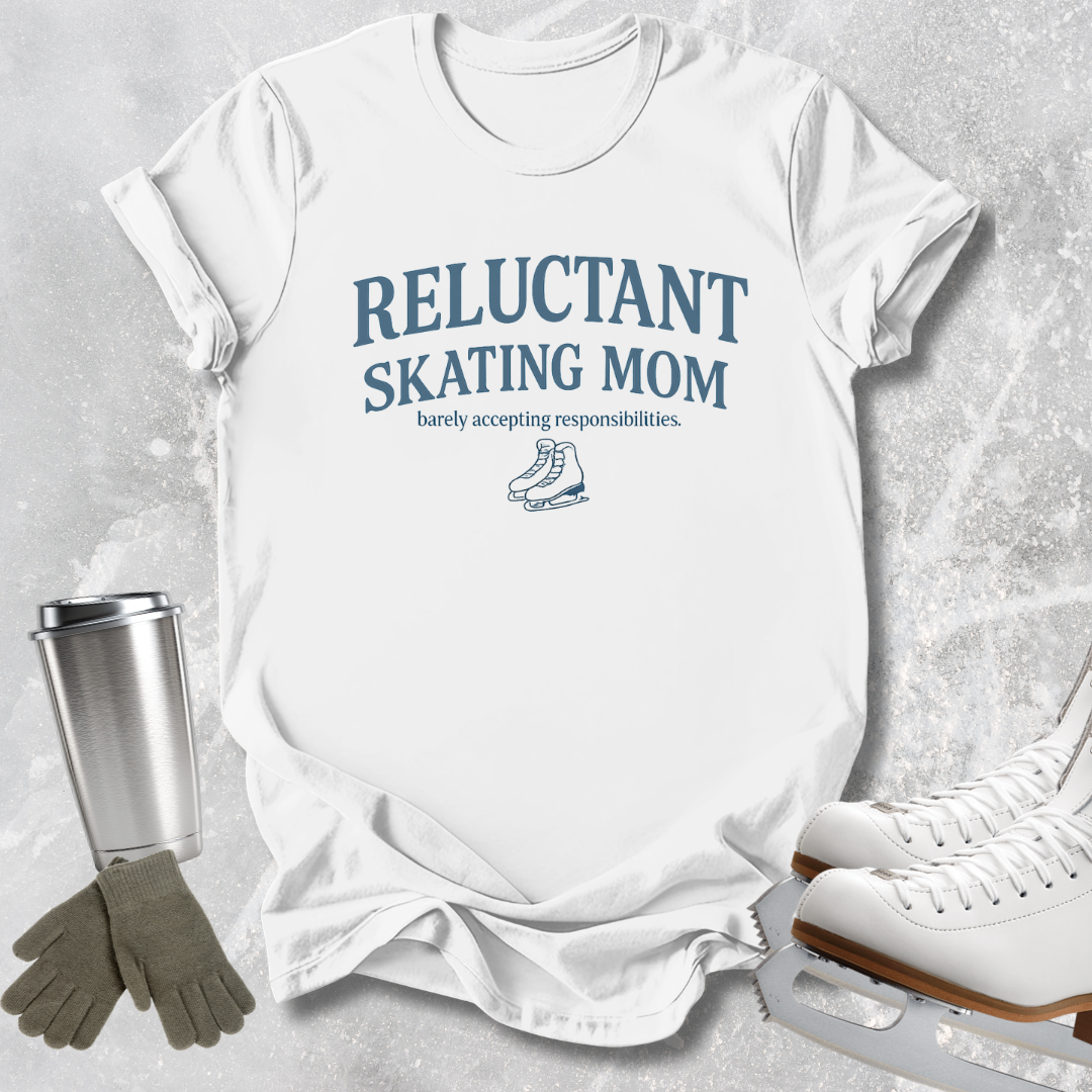 A white short-sleeve T-shirt laid flat on a light gray textured surface, printed with the phrase 'RELUCTANT SKATING MOM' and a small ice skate graphic in blue. Surrounding props include a stainless steel travel mug, gray gloves, and white ice skates.