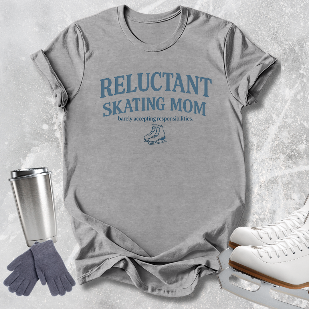 A heather gray short-sleeve t-shirt with a black and pink line-art graphic of a figure skater and the text 'FIGURE SKATER' printed on the front. The shirt is styled in a flat-lay arrangement with ice skates, a stainless steel travel mug, and a pair of gray gloves on a light gray textured background.