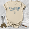 A beige graphic T-shirt laid flat on a light gray textured surface, with blue text reading 'RELUCTANT SKATING MOM' and smaller text below. Ice skates, gloves, and a metal travel mug are styled around the shirt as props.