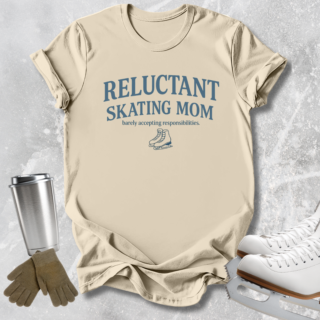 A beige graphic T-shirt laid flat on a light gray textured surface, with blue text reading 'RELUCTANT SKATING MOM' and smaller text below. Ice skates, gloves, and a metal travel mug are styled around the shirt as props.