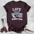 A dark brown graphic t-shirt laid flat on a light gray textured background, styled with ice skates, gloves, and a metal tumbler. The shirt features the text 'LIFE isn't all ABOUT SKATING & DOGS BUT IT SHOULD BE.