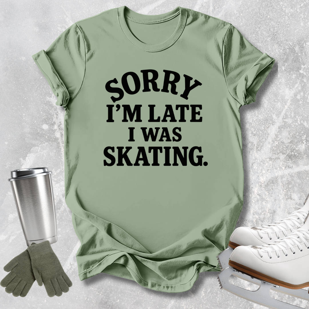 A sage green graphic t-shirt with the text 'SORRY I'M LATE I WAS SKATING.' in black, styled in a flat lay with ice skates, gloves, and a travel tumbler on a textured gray background.