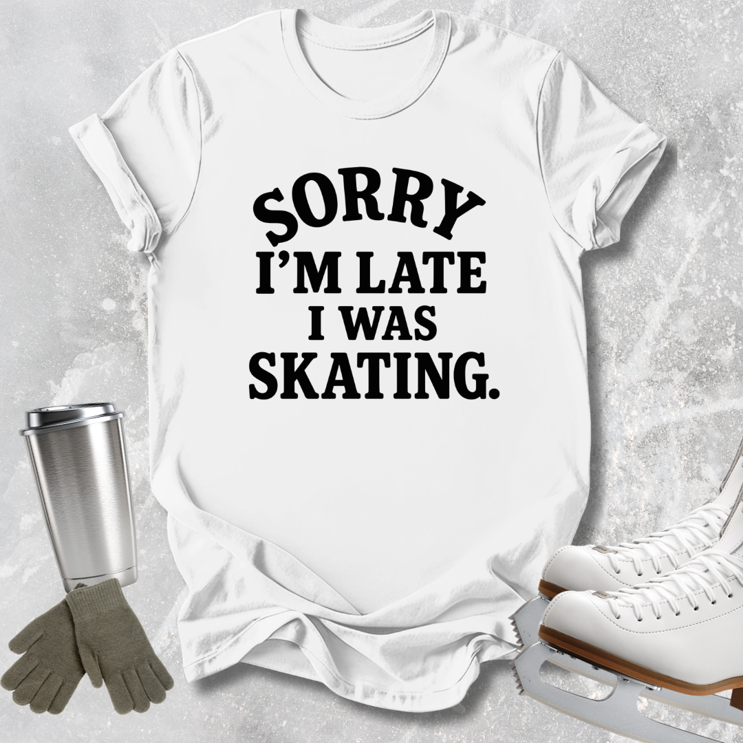 A white short-sleeve T-shirt with bold black text reading 'SORRY I'M LATE I WAS SKATING.' laid flat on a light gray textured surface, styled with ice skates, gloves, and a metal cup nearby.
