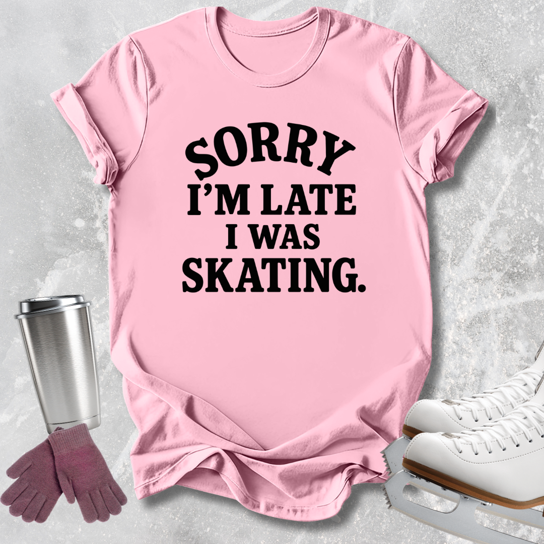 A pink short-sleeve graphic T-shirt laid flat on a light gray textured surface, with black text reading “SORRY I’M LATE I WAS SKATING.” Ice skate shoes, gloves, and a metal cup are arranged around it as styling props.