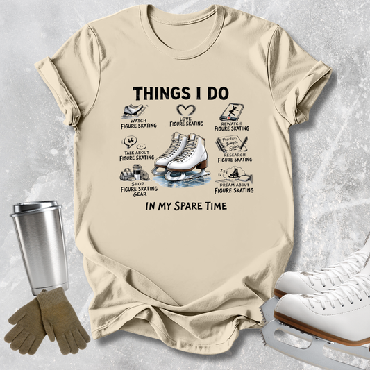 A beige graphic t-shirt featuring a black and white print of ice skates and text reading 'THINGS I DO IN MY SPARE TIME' with small illustrations and labels. The t-shirt is displayed in a flat-lay arrangement on a textured gray background, surrounded by ice skating props including white ice skates, a metal travel tumbler, and a pair of brown gloves.