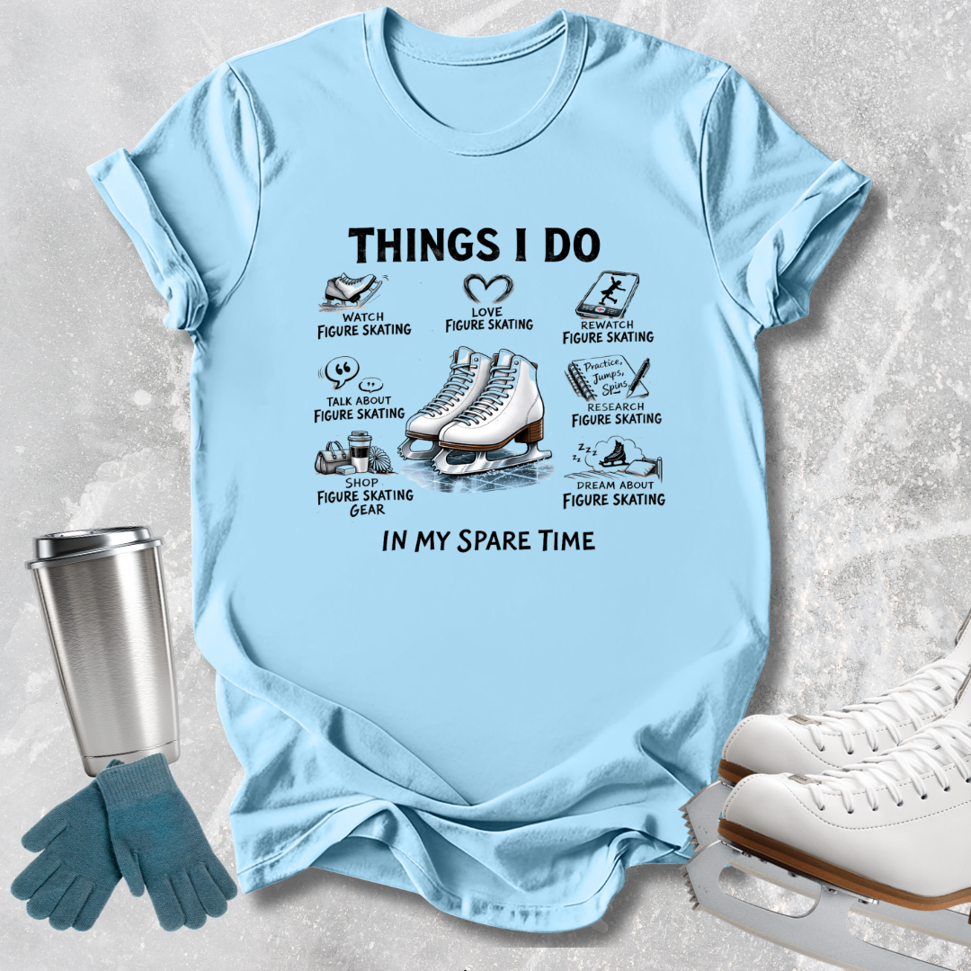 A light blue graphic t-shirt featuring a black and white print of ice skates and text reading 'THINGS I DO IN MY SPARE TIME' with various figure skating activities. The shirt is displayed in a flat-lay arrangement with ice skates, gloves, and a travel tumbler on a textured gray background.
