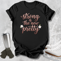 A black short-sleeve graphic T-shirt laid flat on a light stone-textured background, with the phrase "strong is the new pretty" in rose-gold script and two small white flowers. White ice skates, dark gloves, and a stainless steel travel tumbler are arranged around the shirt as styling props.