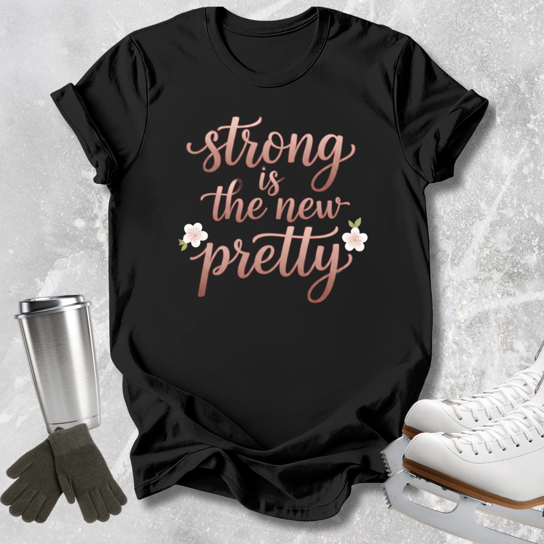A black short-sleeve graphic T-shirt laid flat on a light stone-textured background, with the phrase "strong is the new pretty" in rose-gold script and two small white flowers. White ice skates, dark gloves, and a stainless steel travel tumbler are arranged around the shirt as styling props.
