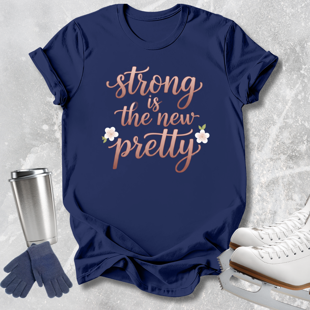A navy blue graphic T-shirt laid flat on a light stone-textured background, with the phrase "strong is the new pretty" in rose-gold script and two small white flowers. Surrounding props include a stainless steel travel tumbler, navy gloves, and white ice skates.