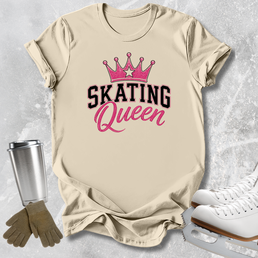 A beige graphic T-shirt laid flat on a light gray textured surface, featuring a pink crown and the words 'SKATING Queen' in black and pink. Ice skates, gloves, and a metal tumbler are styled around the shirt as props.