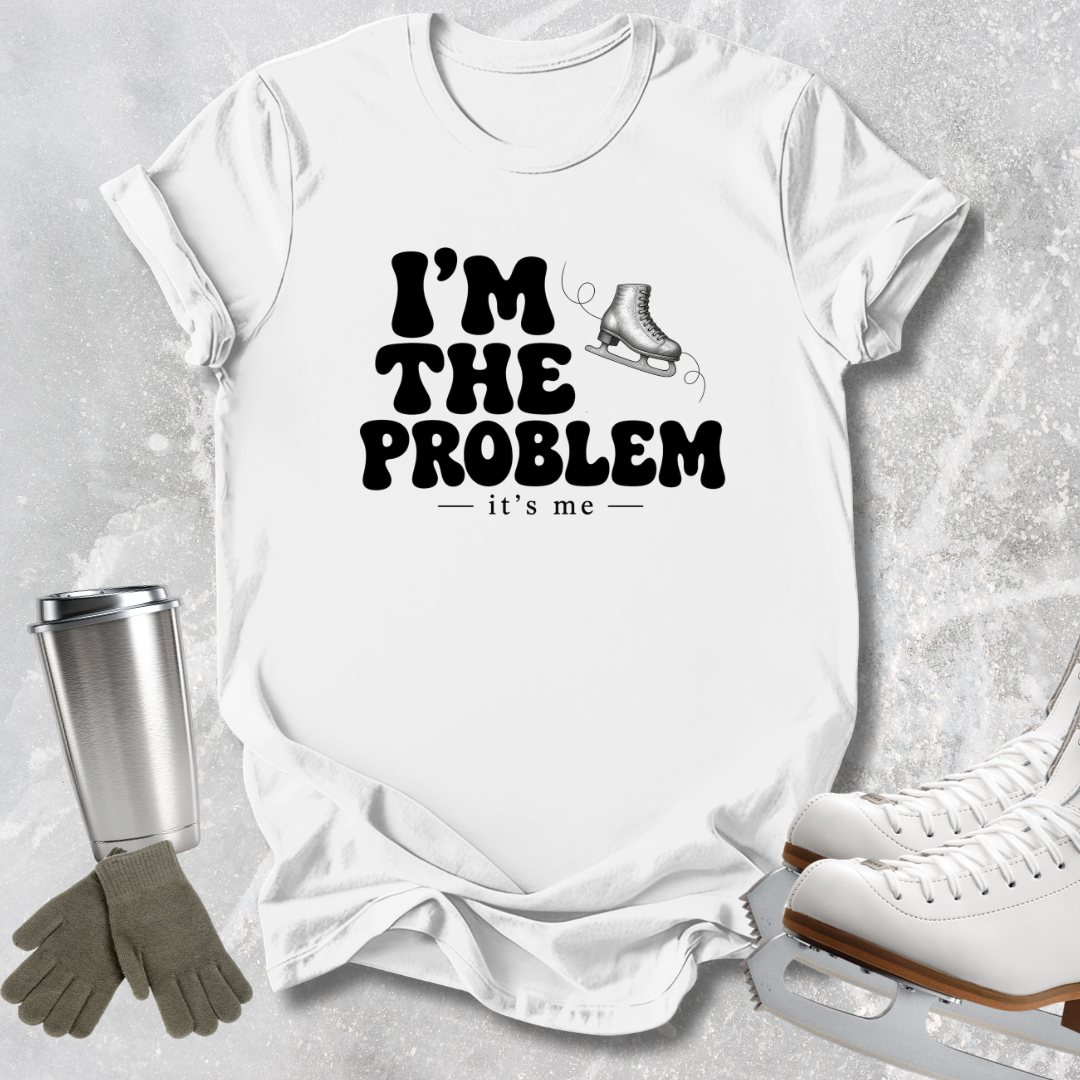 A white short-sleeve graphic T-shirt laid flat on a light gray textured surface, with black text reading “I’M THE PROBLEM it’s me” and a small ice skate illustration. Ice skates, gloves, and a metal cup are placed around the shirt as styled props.