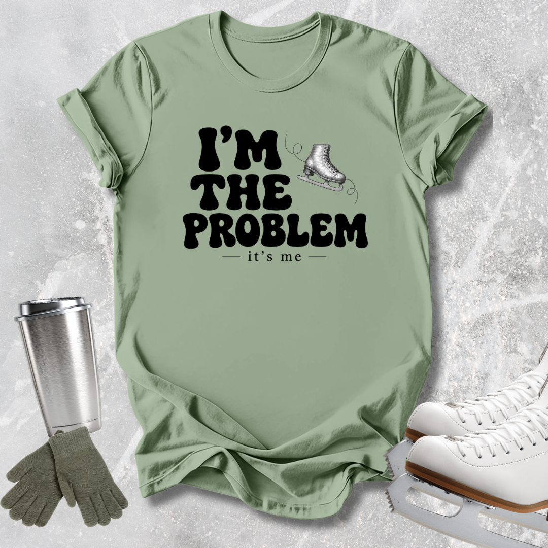 A sage green graphic t-shirt laid flat on a textured gray background, styled with ice skates, a travel tumbler, and gloves. The t-shirt features the text 'I'M THE PROBLEM - it's me' in black with a small ice skate graphic.