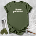 An olive green graphic t-shirt with the text 'i have potential' in white, styled in a flat-lay with ice skates, a travel tumbler, and gloves on a textured gray background.