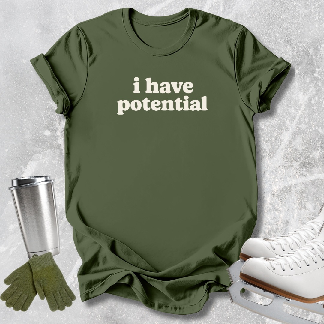 An olive green graphic t-shirt with the text 'i have potential' in white, styled in a flat-lay with ice skates, a travel tumbler, and gloves on a textured gray background.