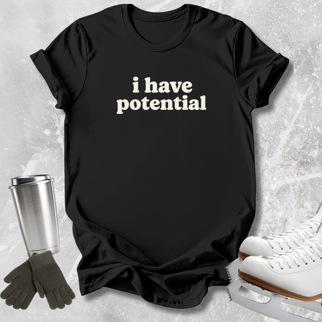 A black short-sleeve T-shirt with the phrase 'i have potential' printed in white serif text across the chest, styled in a flat lay with white ice skates, dark gloves, and a stainless steel tumbler on a light gray textured background.