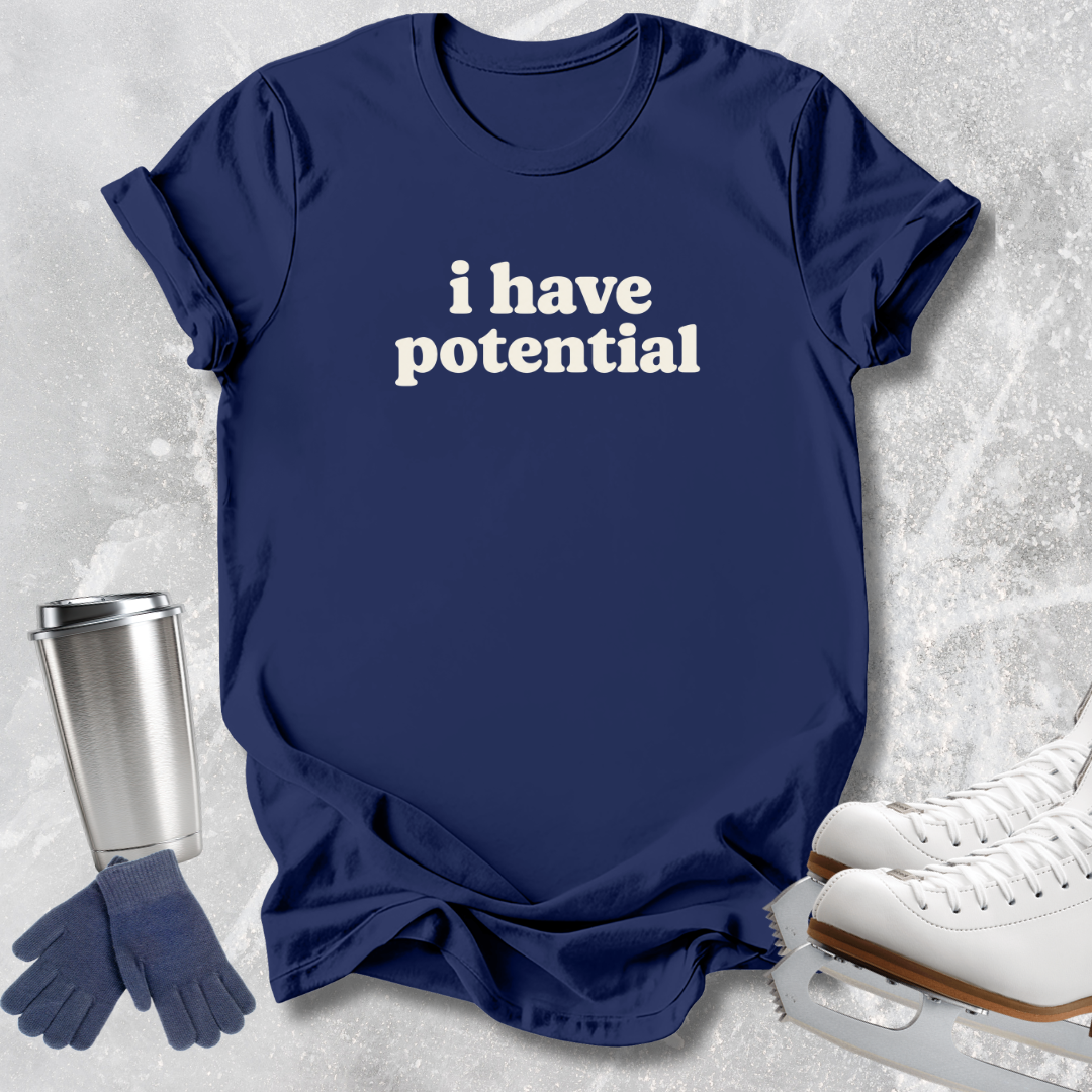 A navy blue graphic t-shirt with the phrase 'i have potential' printed in white text across the chest, styled in a flat-lay arrangement with ice skates, gloves, and a metal tumbler on a light gray textured background.