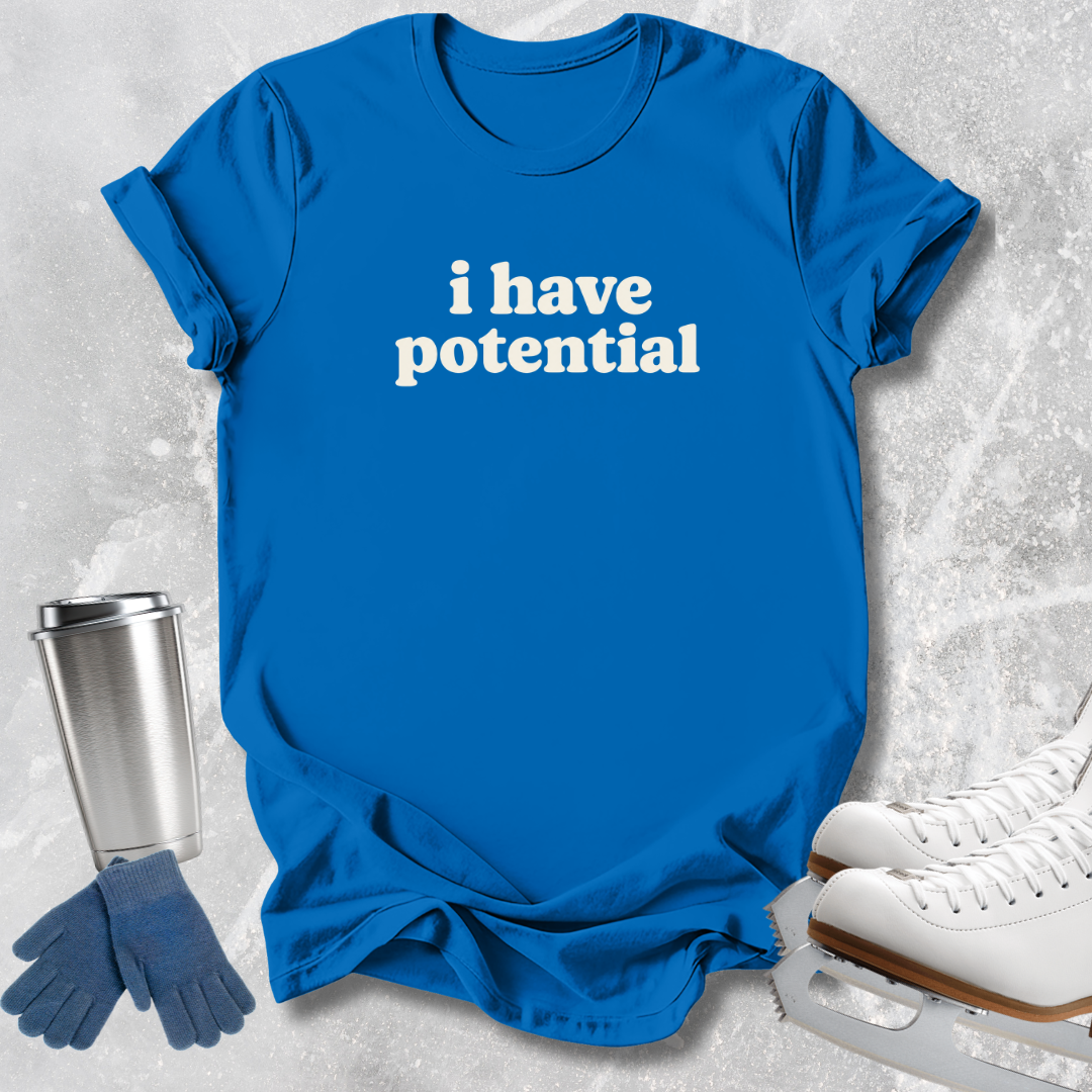 A bright blue short-sleeve T-shirt with the phrase 'i have potential' printed in white across the chest, styled in a flat lay with ice skates, gloves, and a metal travel tumbler on a light gray textured surface.