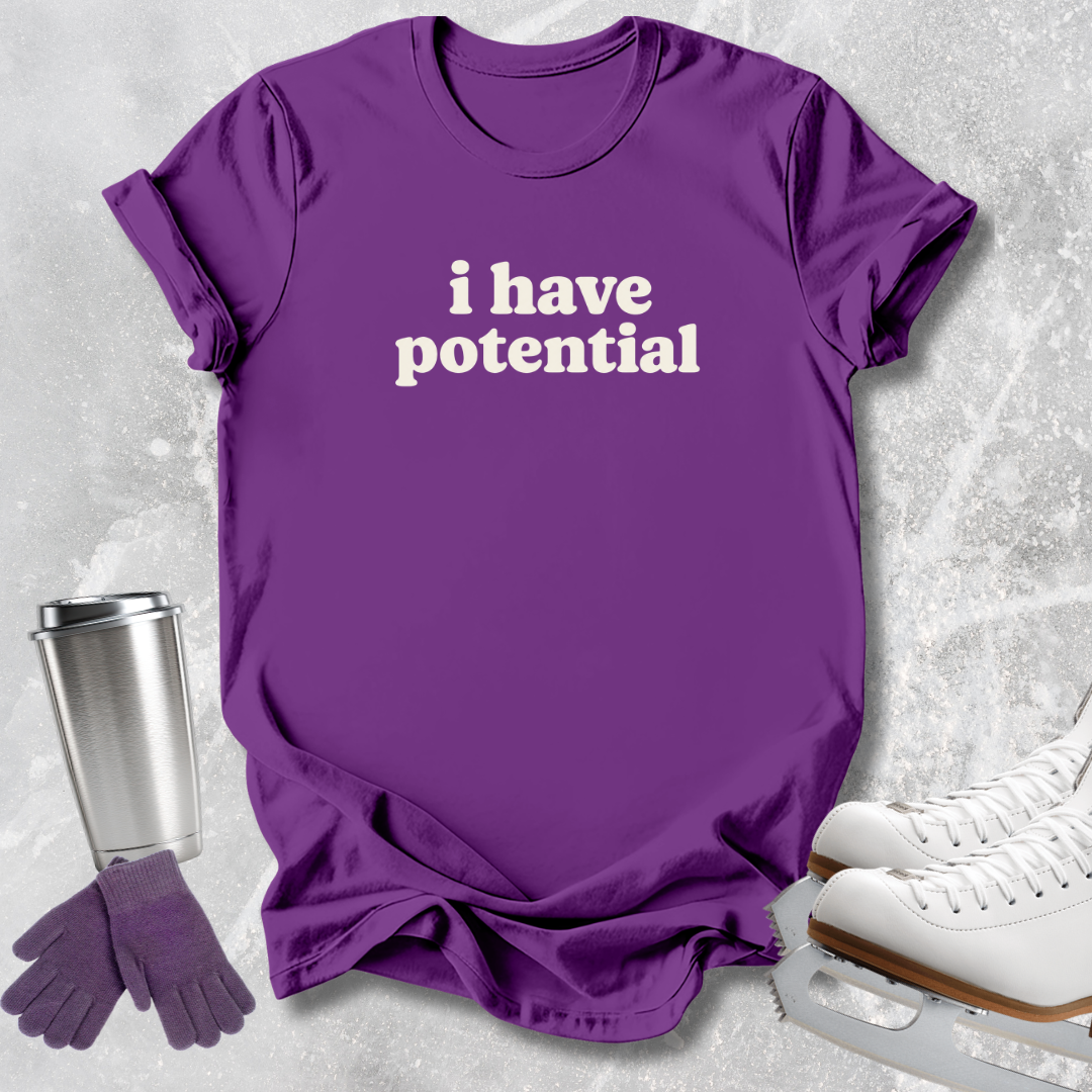 A purple short-sleeve T-shirt with the phrase 'i have potential' printed in white across the chest, styled in a flat lay with ice skates, gloves, and a travel tumbler on a light gray textured background.