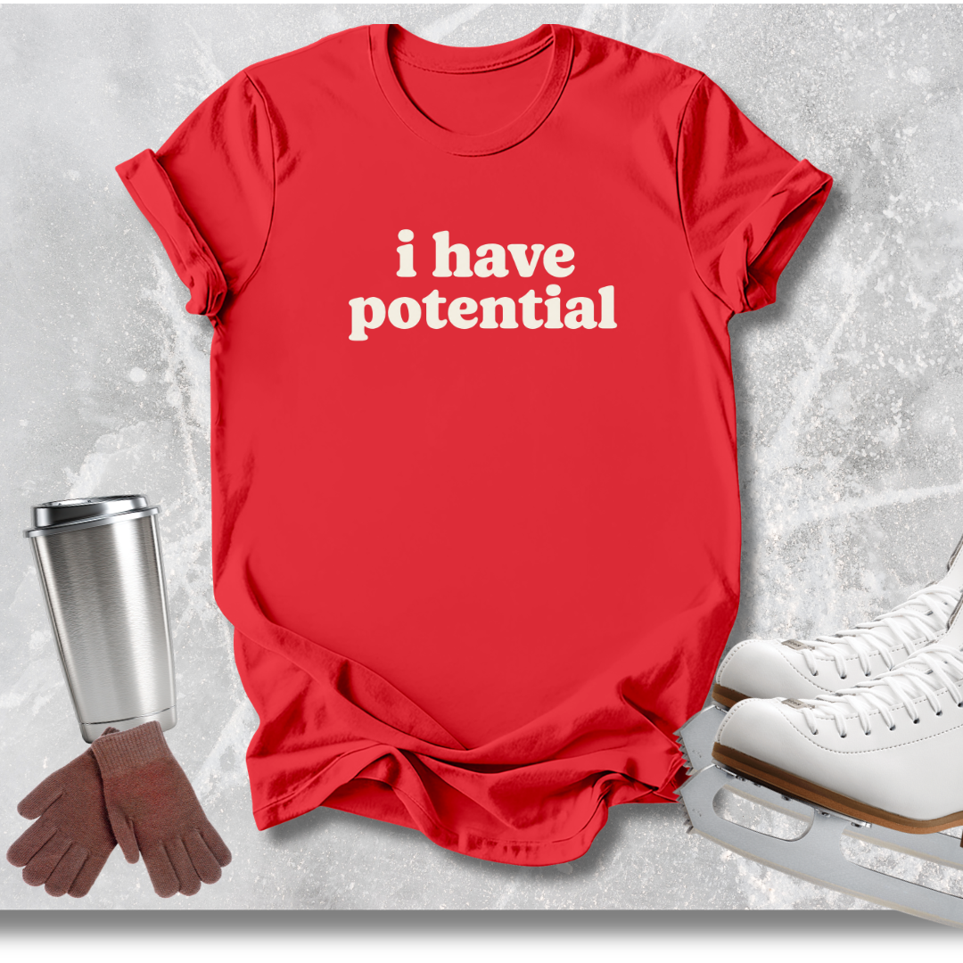 A red short-sleeve T-shirt with the phrase 'i have potential' printed in white across the chest, styled in a flat lay with ice skates, gloves, and a metal cup on a light gray textured background.