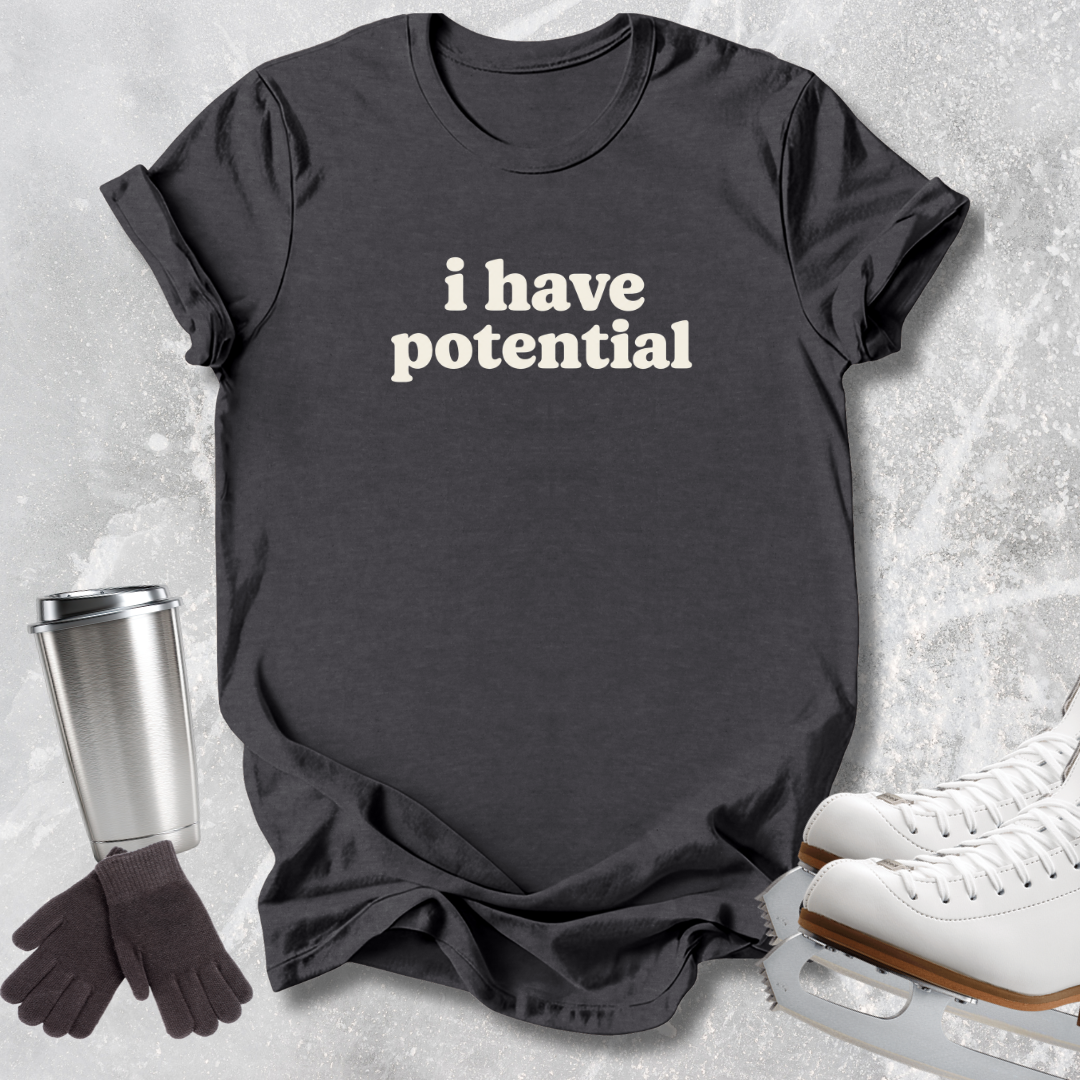 A dark charcoal gray graphic t-shirt with the text 'i have potential' printed in white lowercase letters. The t-shirt is styled in a flat-lay arrangement on a light gray textured background, accompanied by a stainless steel travel mug, a pair of black gloves, and white ice skates.