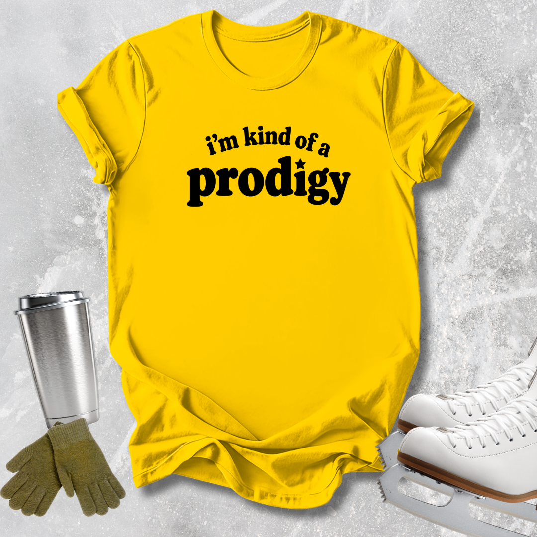 A bright yellow graphic t-shirt with the text 'i'm kind of a prodigy' printed in black across the chest, styled in a flat-lay with ice skates, gloves, and a travel tumbler on a textured gray background.