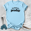 A light blue graphic t-shirt with the text 'i'm kind of a prodigy' printed in black across the chest, styled in a flat-lay with ice skating props including white ice skates, blue gloves, and a stainless steel tumbler on a textured gray background.