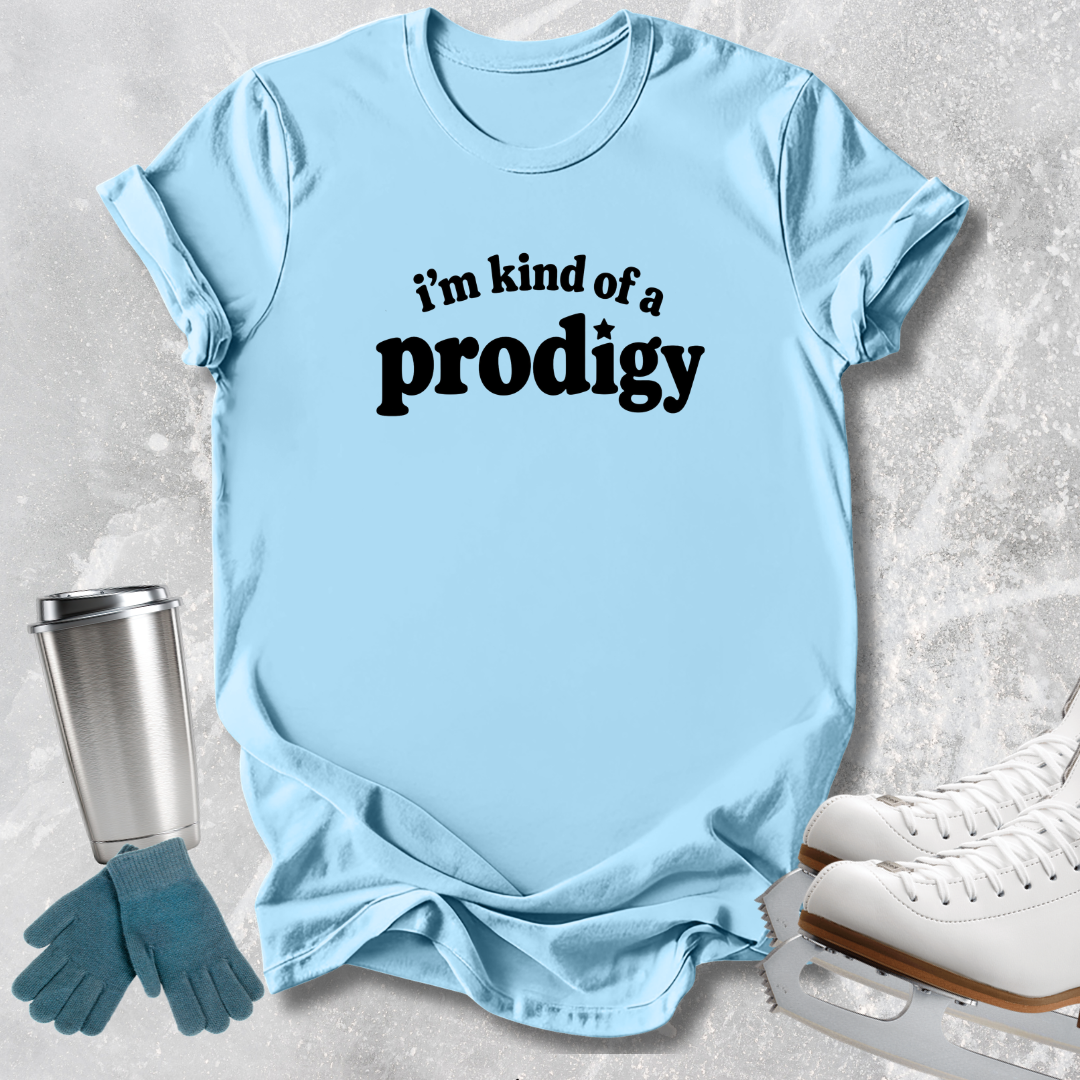 A light blue graphic t-shirt with the text 'i'm kind of a prodigy' printed in black across the chest, styled in a flat-lay with ice skating props including white ice skates, blue gloves, and a stainless steel tumbler on a textured gray background.