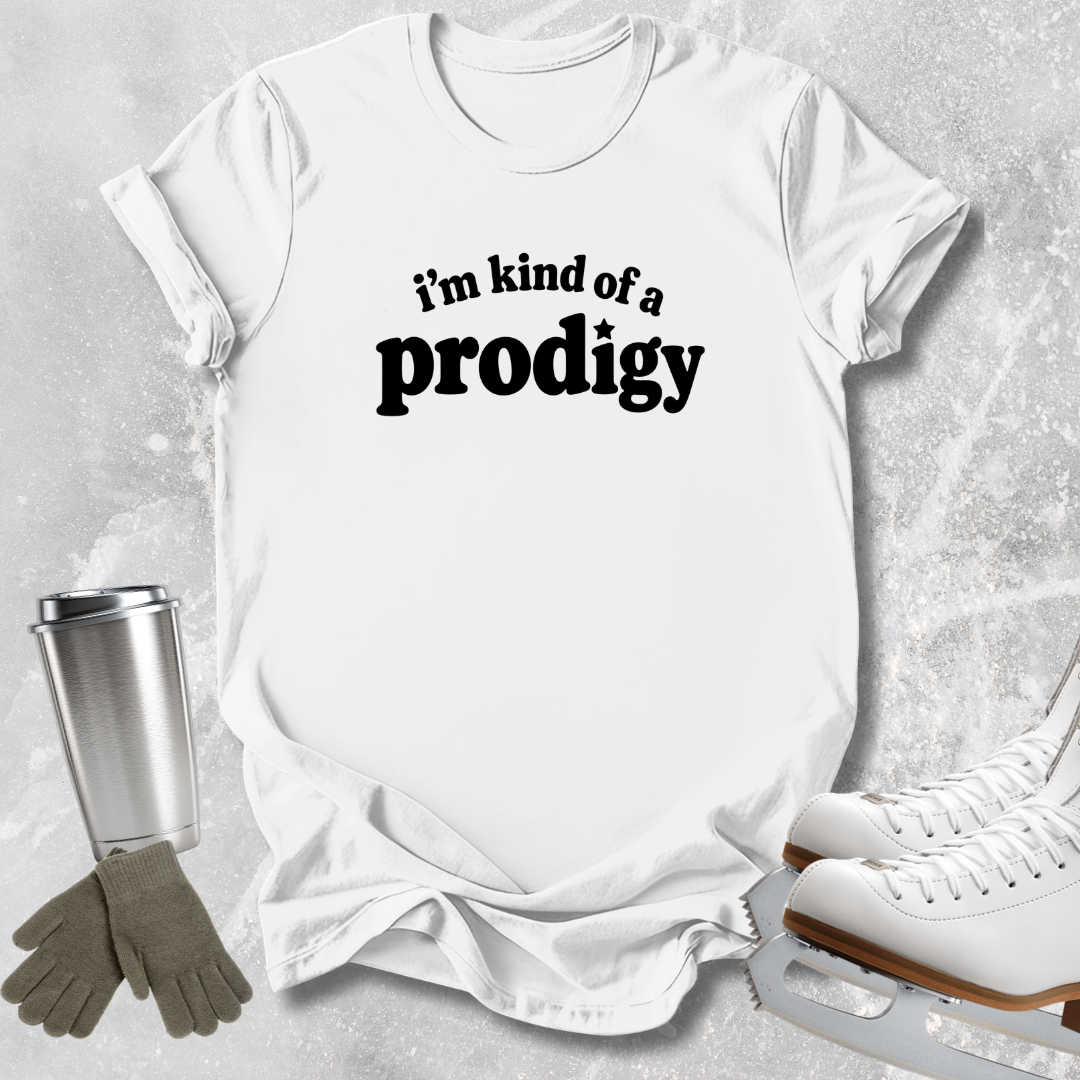 A white short-sleeve graphic T-shirt laid flat on a light gray textured surface, with the phrase "i'm kind of a prodigy" printed in black across the chest. Ice skates, gloves, and a metal tumbler are arranged around it as styling props.
