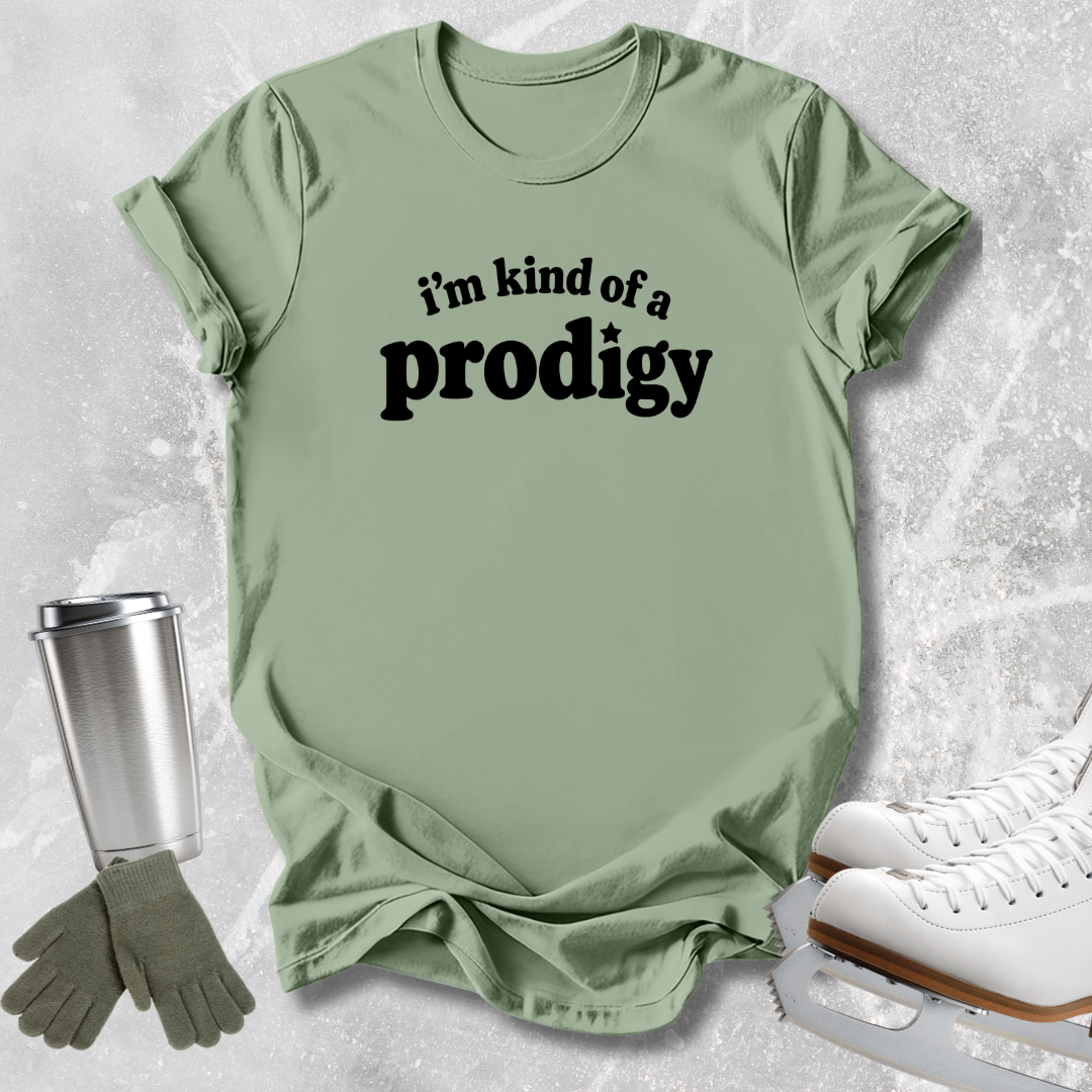 A sage green graphic t-shirt with the text 'i'm kind of a prodigy' printed in black across the chest, styled in a flat lay with ice skates, a travel tumbler, and gloves on a light gray textured background.