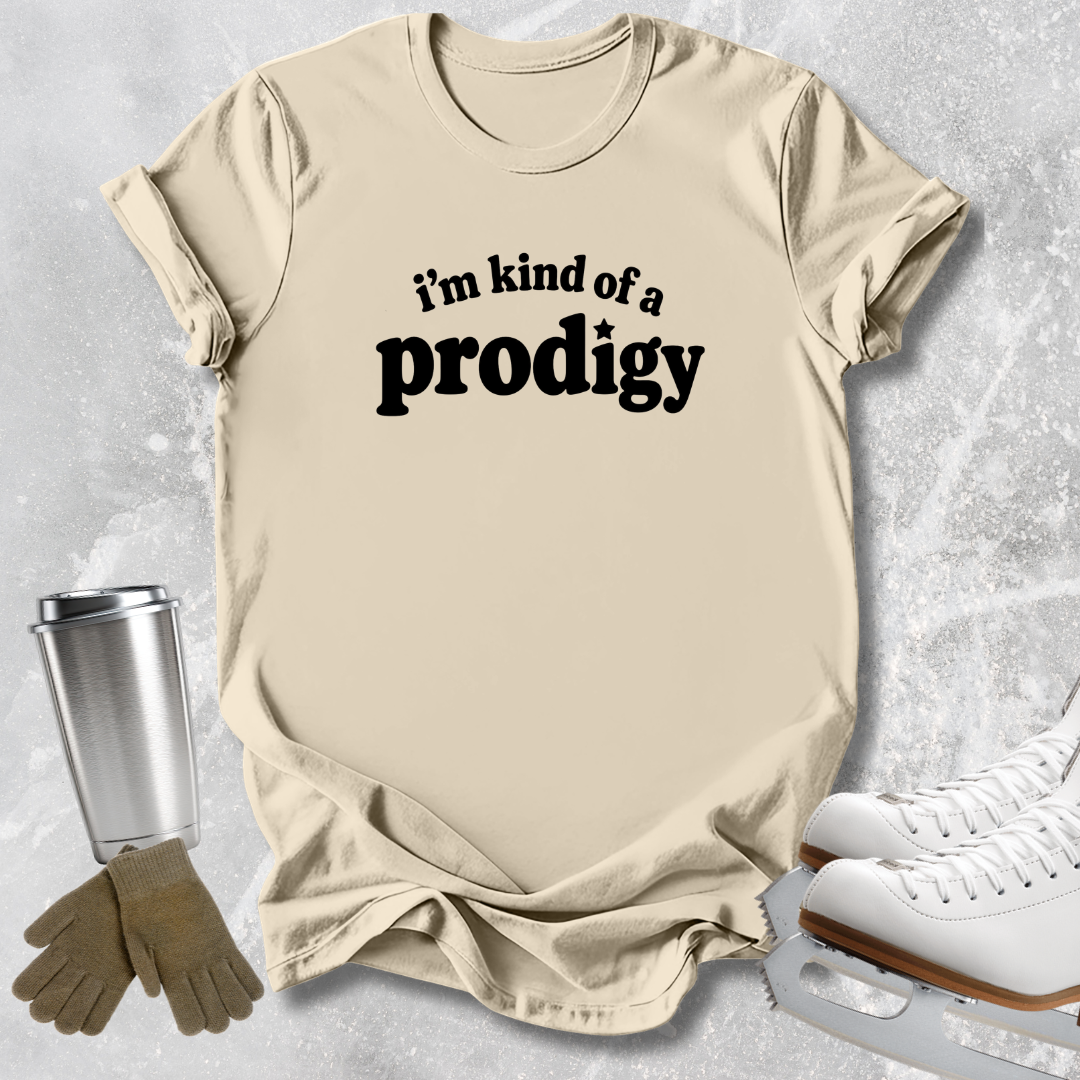 A beige graphic t-shirt with the text 'i'm kind of a prodigy' in black, styled in a flat-lay with ice skates, gloves, and a travel tumbler on a light gray textured background.